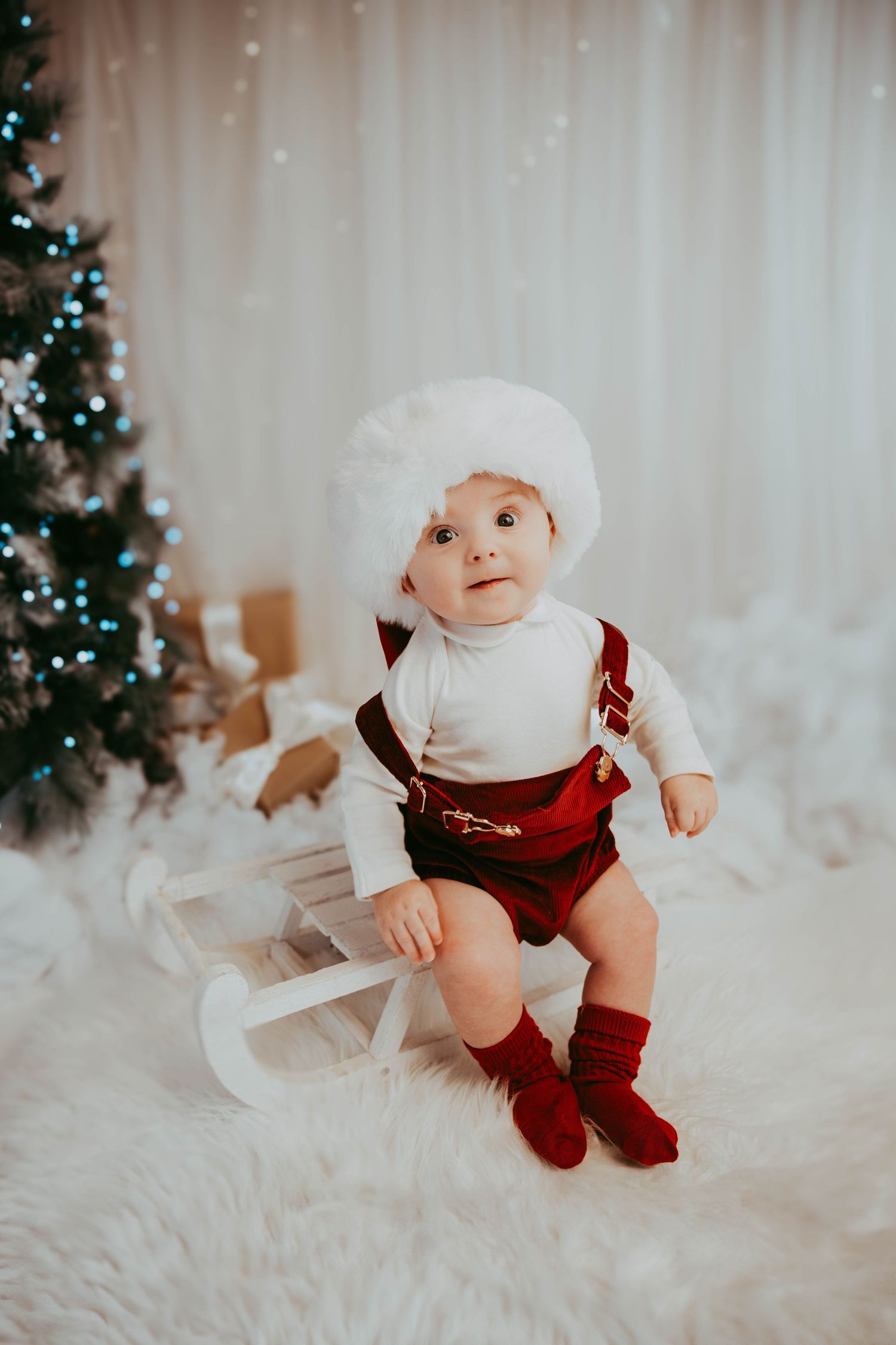 Baby in red velvet overalls and white fur hat sits on small sleigh with Christmas decor.