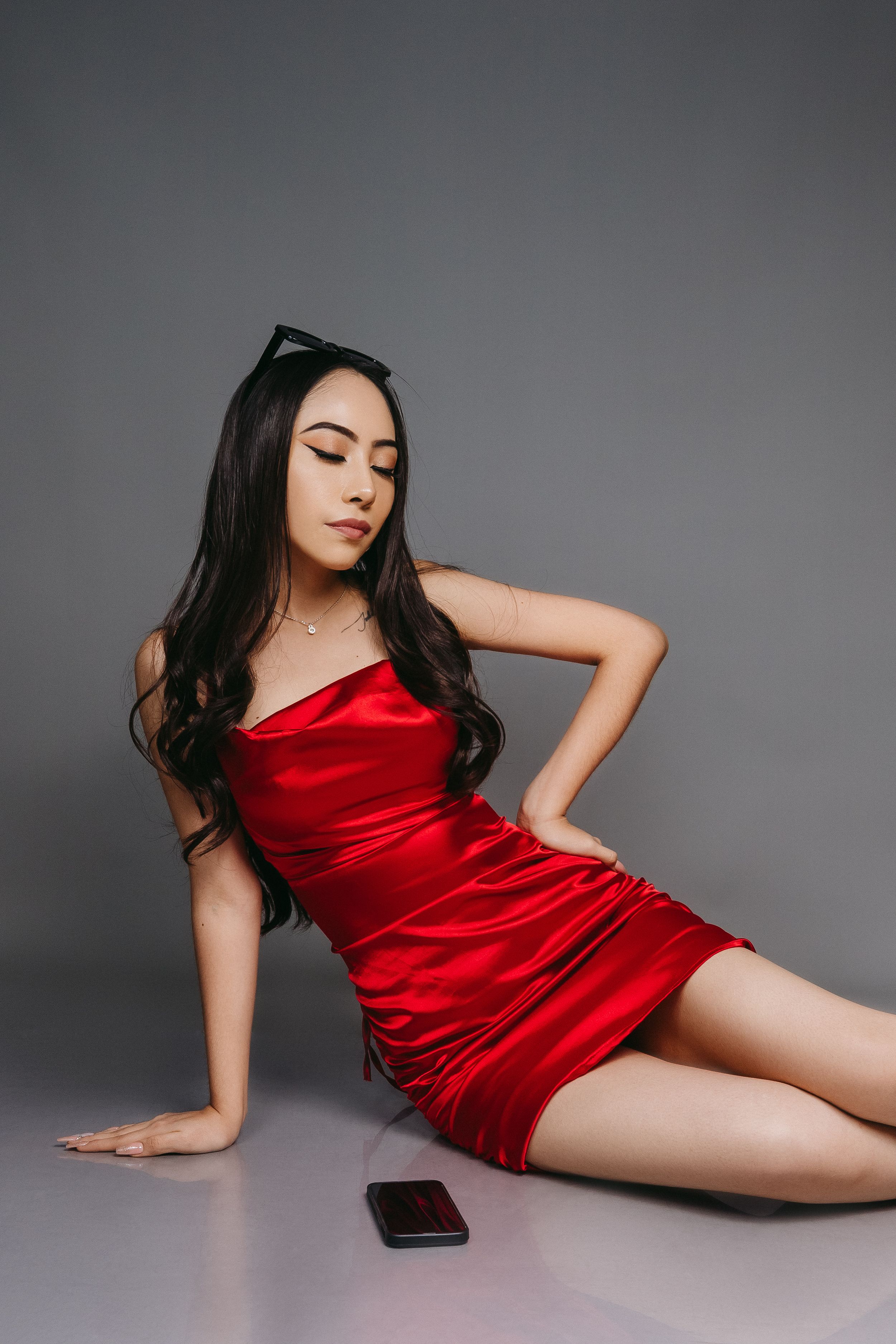 Model in red satin mini dress poses on floor with phone nearby against grey backdrop.