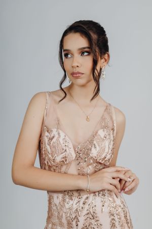 Model in rose gold sequined gown with sheer details poses elegantly against grey background.