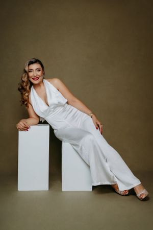Elegant model in white halter jumpsuit poses on geometric white blocks against brown backdrop.