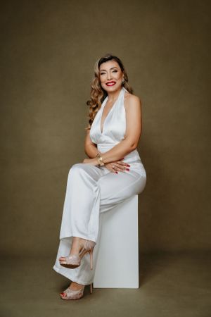 Elegant model in white jumpsuit sits gracefully on cube with retro-styled hair and makeup.