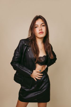 Fashion model poses in edgy black leather jacket and skirt outfit against beige background.