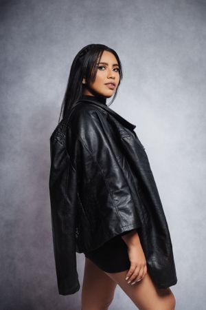 Model wearing oversized leather jacket poses dramatically against grey studio background.
