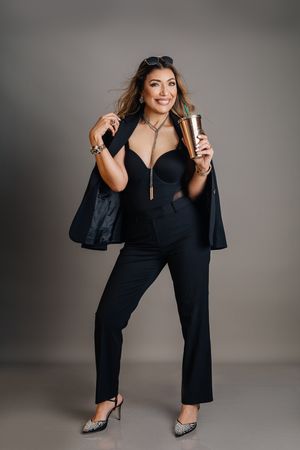Model in black pantsuit and sparkly heels holds metallic tumbler against grey studio backdrop.