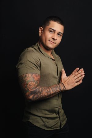 Man in olive shirt shows tattoo sleeve while posing against dark background.