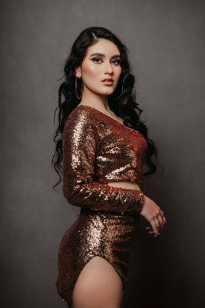 Fashion model in a shimmering bronze sequin dress with long dark wavy hair poses against grey backdrop.