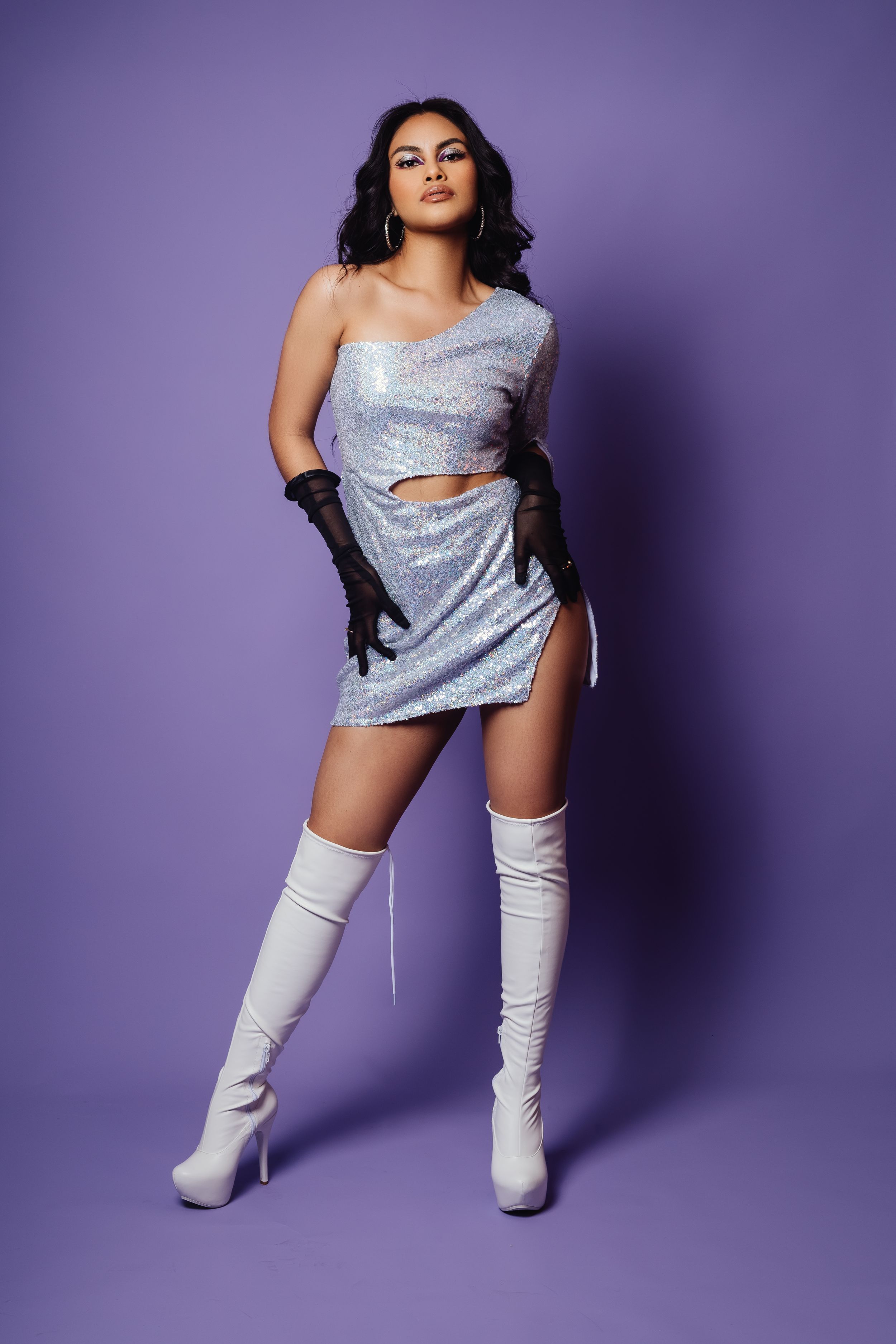 Model wearing silver sequin dress and white boots poses confidently against purple backdrop.