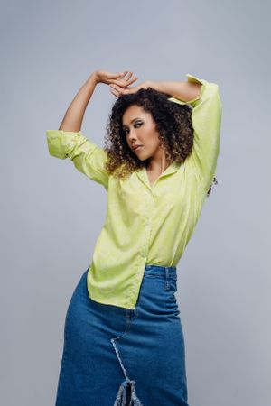 Model in yellow shirt and denim skirt strikes dynamic pose on grey backdrop.