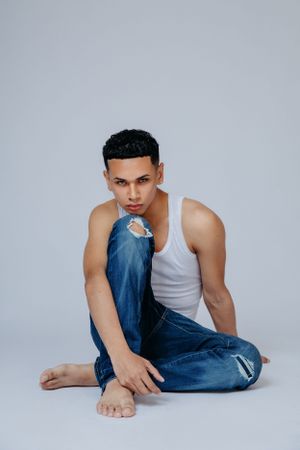 Athletic model in white tank top and distressed jeans poses casually on floor.