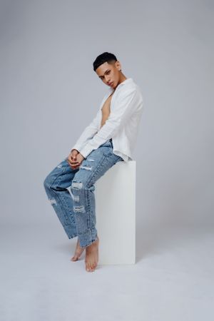 Model in unbuttoned white shirt and ripped jeans sits barefoot on white pedestal.