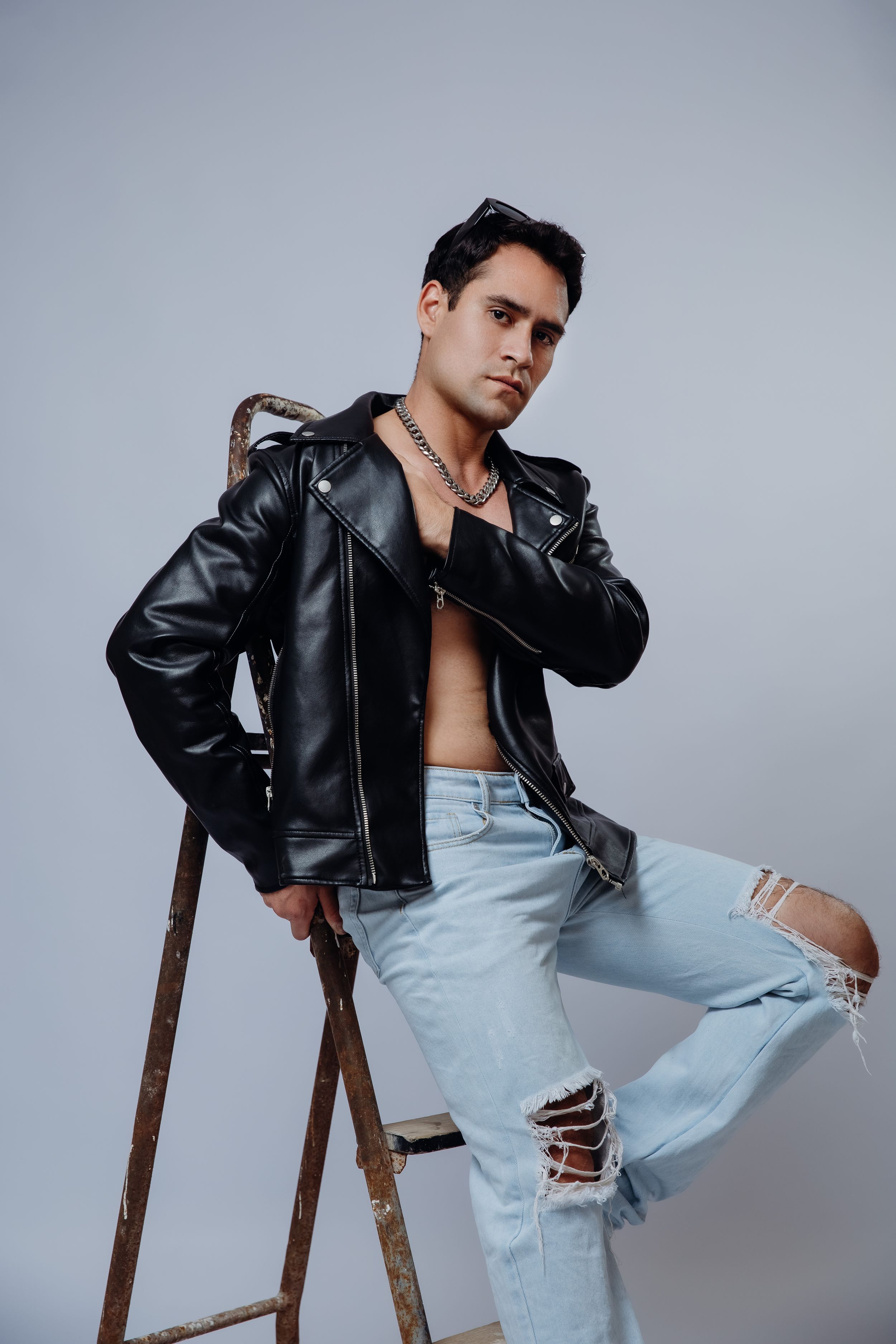 Model wearing leather jacket and ripped jeans poses on wooden stool against grey backdrop.