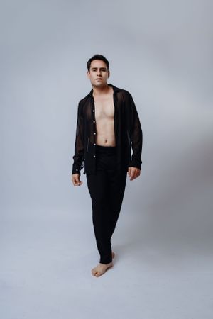 Model in black open shirt and pants poses against light grey background.
