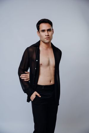 Model in sheer black shirt and dark pants poses in studio setting.