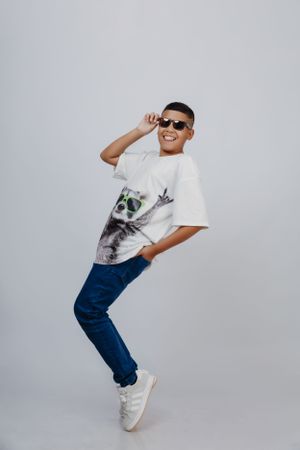 Playful pose in white graphic t-shirt, jeans and sunglasses against light background.
