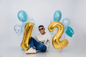 Casual seated pose with gold number 12 balloons and blue party decorations.