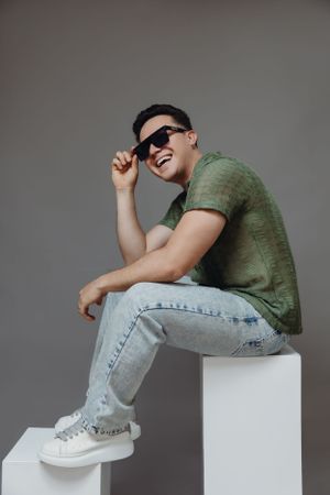 Casual portrait in green t-shirt and jeans sitting on white cube wearing sunglasses.