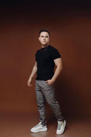 Studio portrait in black t-shirt and grey pants against brown background.