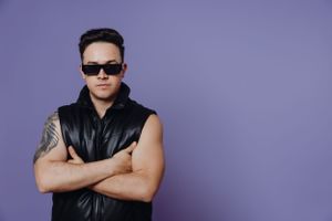 Cool stance in black leather vest and sunglasses against purple background.