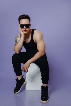 Fashion model in black tank top and sunglasses poses on white cube against purple backdrop.