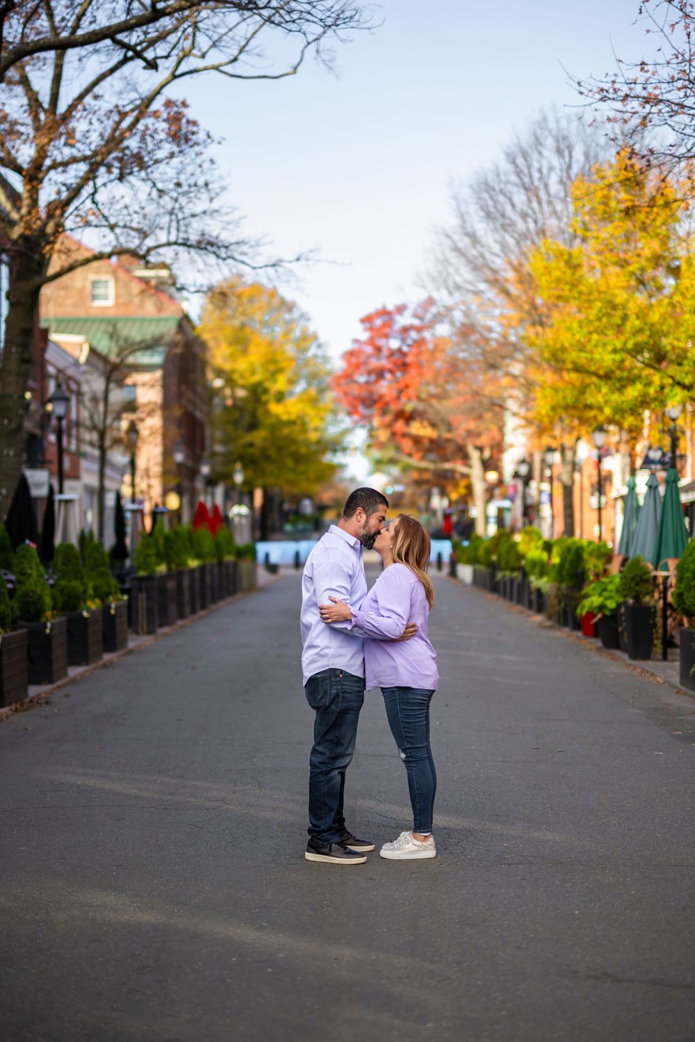 Couple sharing a kiss on street with autumn trees and historic buildings in background.