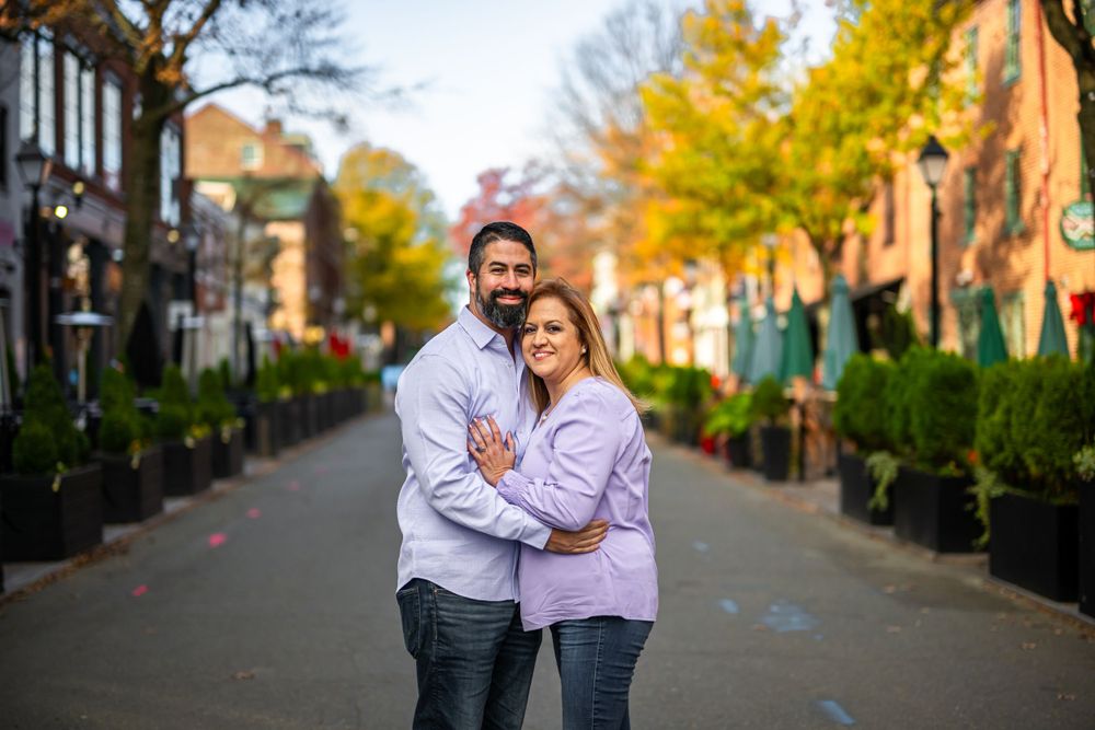 Couple embracing on charming city street lined with autumn trees and potted plants.