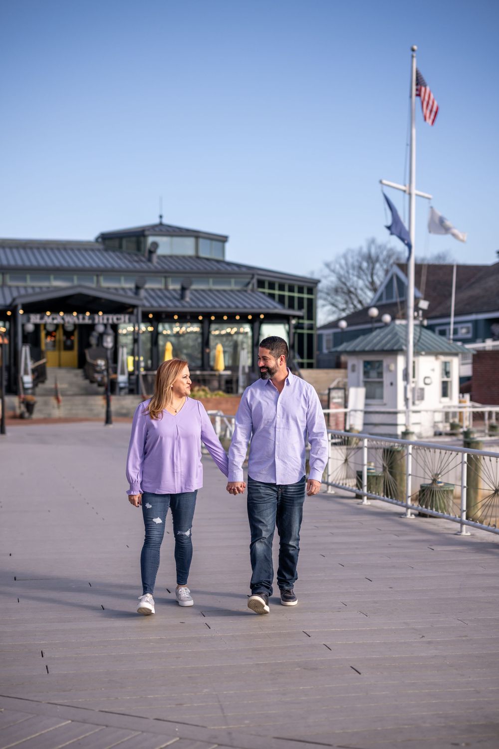 Couple walking hand in hand along waterfront boardwalk with American flag and shopping area.