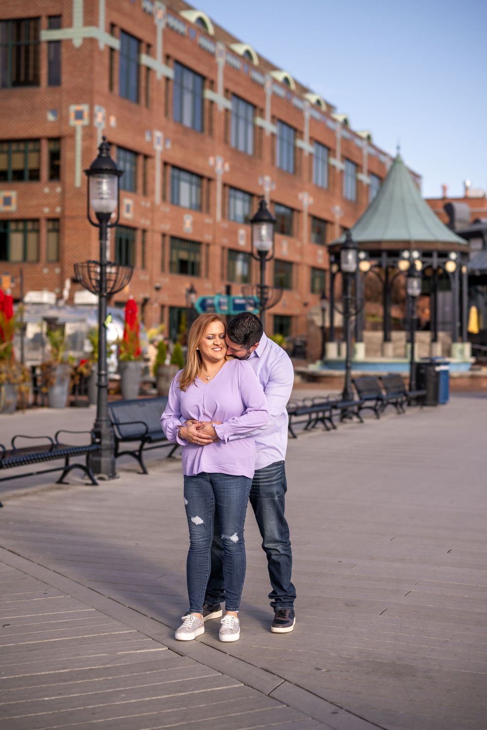 Couple embracing on brick-lined boardwalk with historic building and lampposts in background.