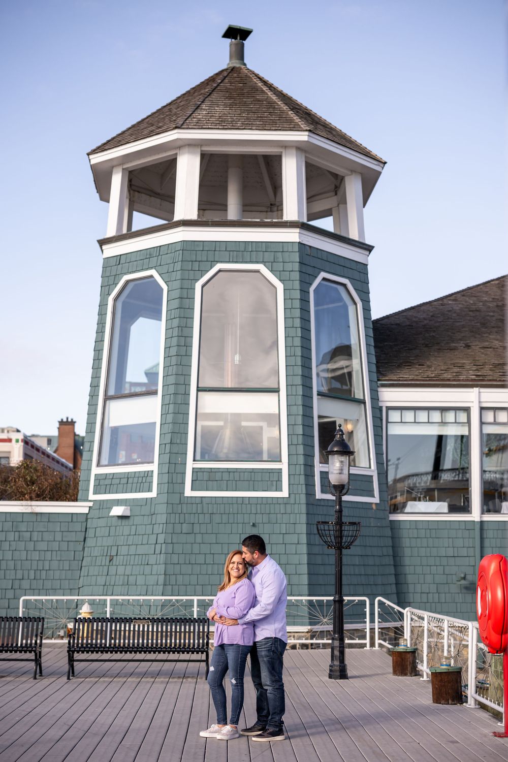 Couple standing close together by coastal tower with green shingles and viewing deck.
