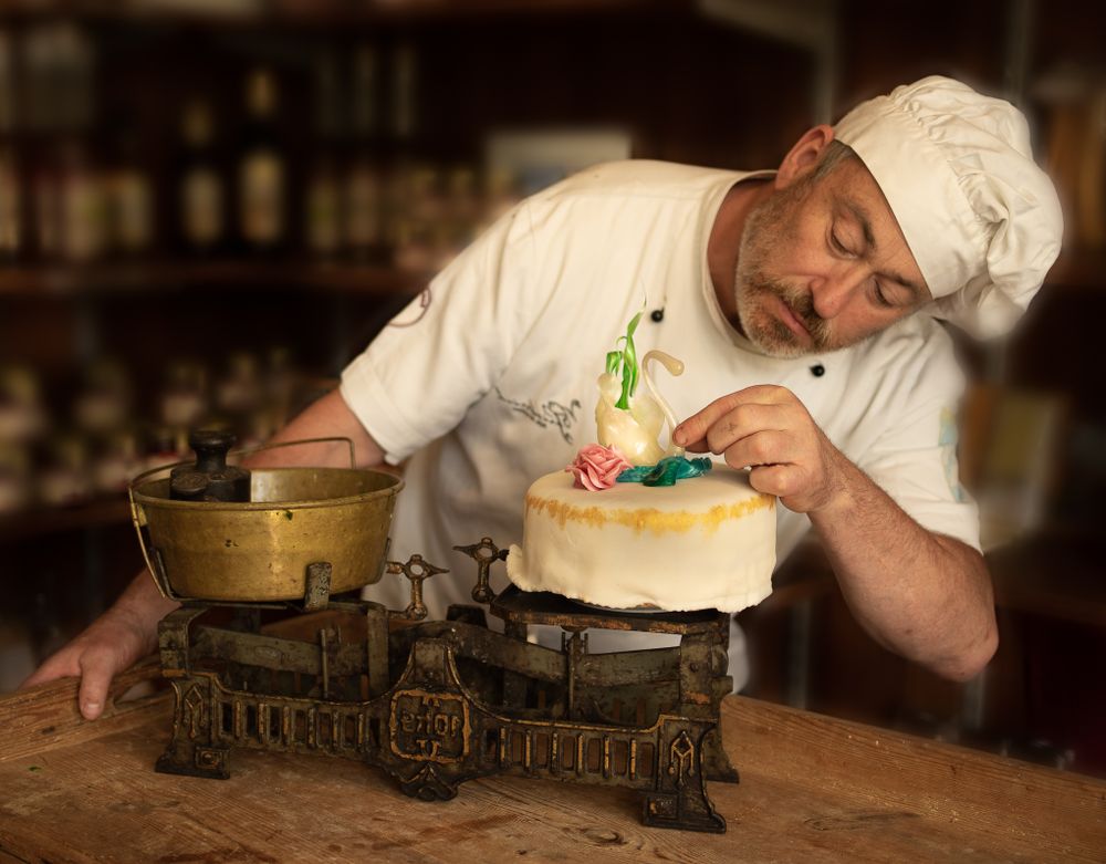 BusinessFotografie, Business Projects. Chef in white uniform carefully decorates a cake on vintage scale with delicate garnishes.