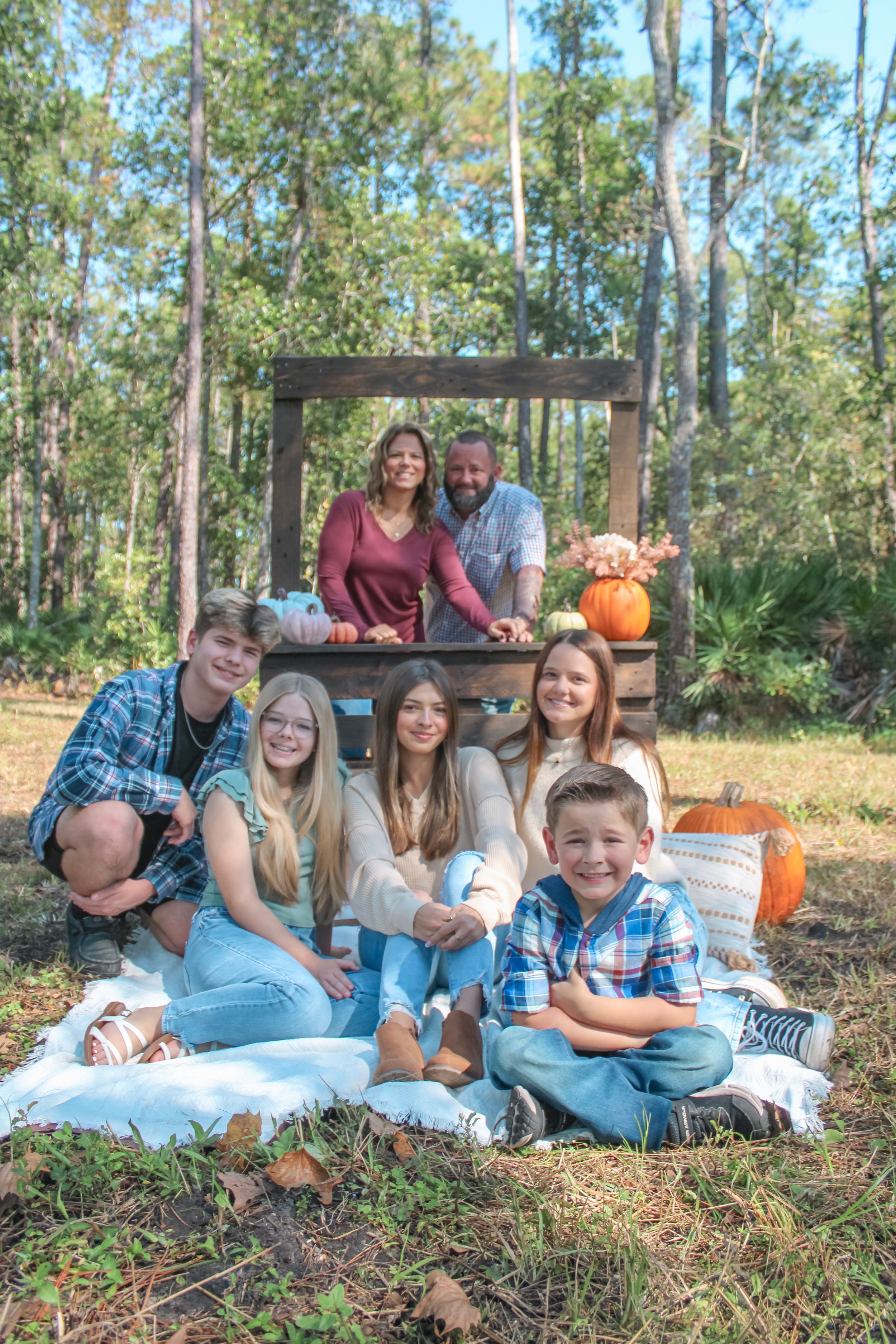 Family gathering outdoors with pumpkins and blankets for autumn photo session among pine trees.
