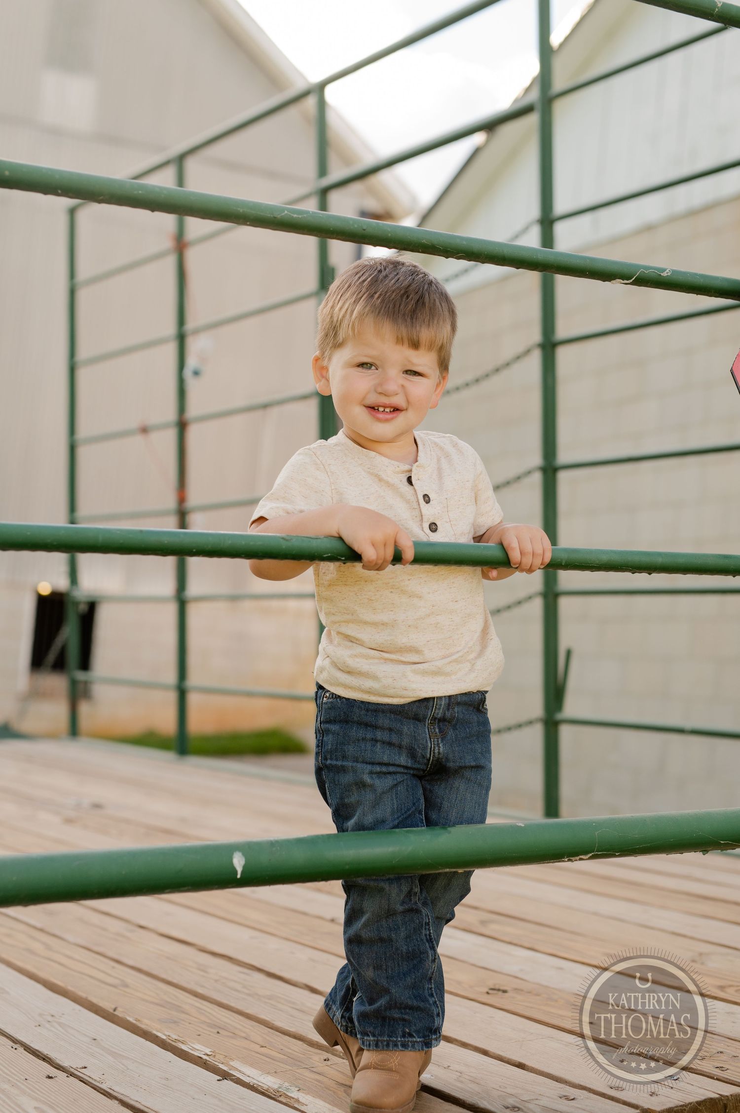 Young child in a white shirt and jeans plays on green metal railings with joyful expression.