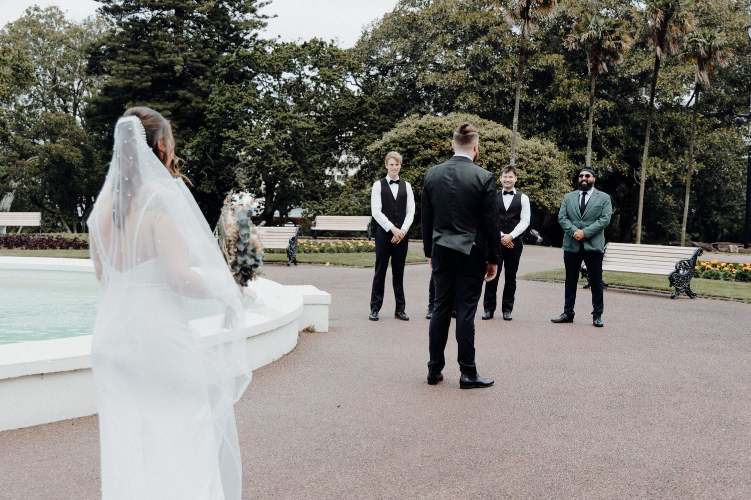 A bride in a flowing white veil stands near a fountain for the First Look, captured by Apertured Media Auckland Wedding Photographer.