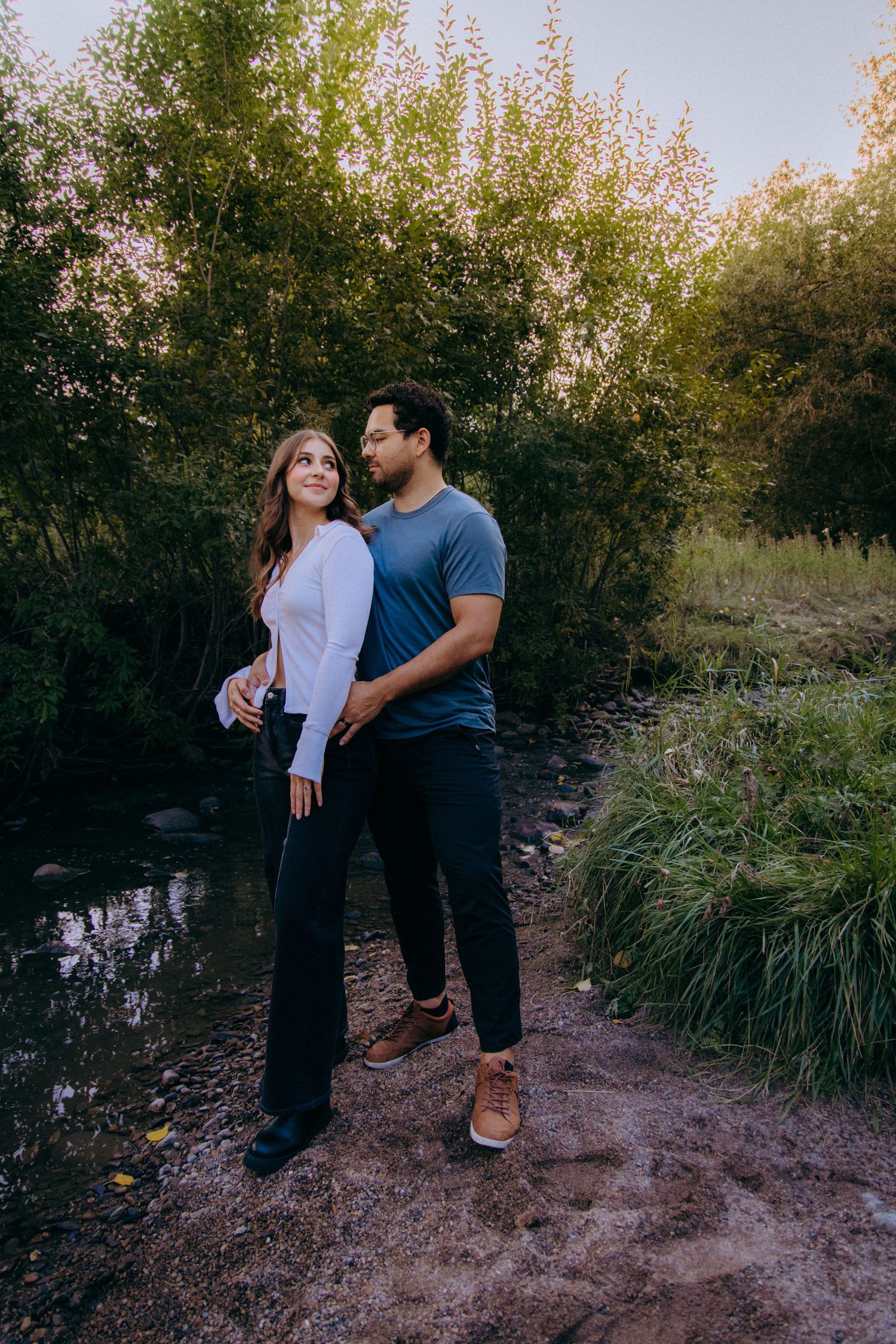 A couple shares an intimate moment by a pond surrounded by tall grass and trees at sunset.