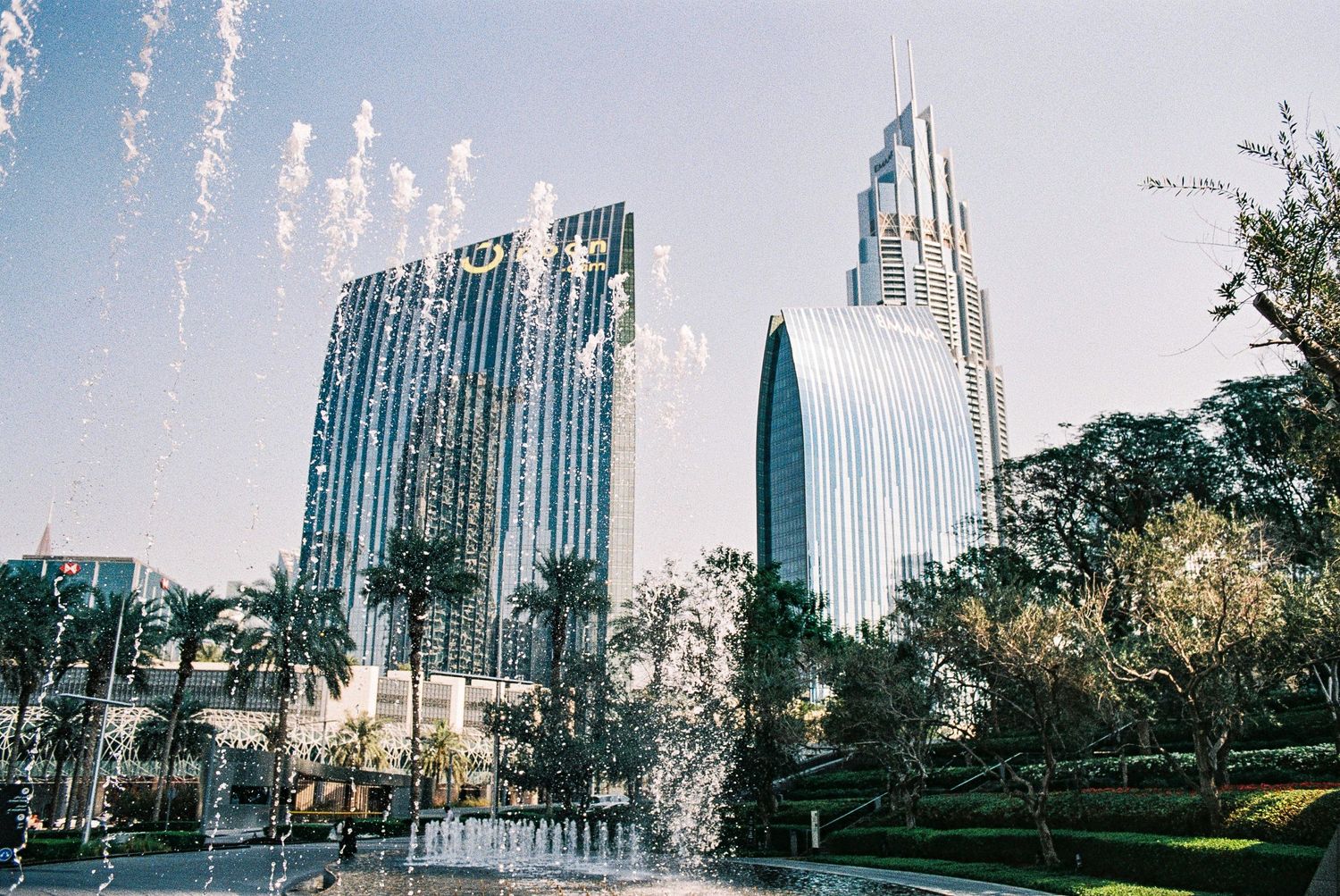 Modern glass skyscrapers and palm trees in Dubai's downtown area reflect sunlight against a bright blue sky.