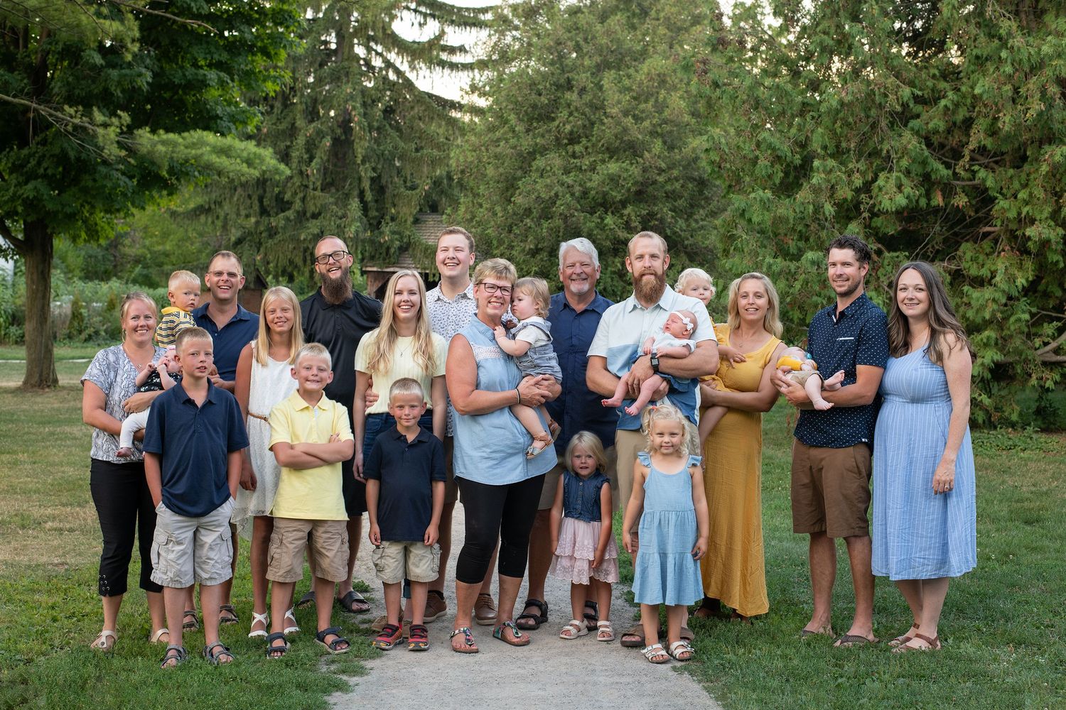 A large multi-generational group gathers outdoors on a summer evening for a portrait in a park setting.