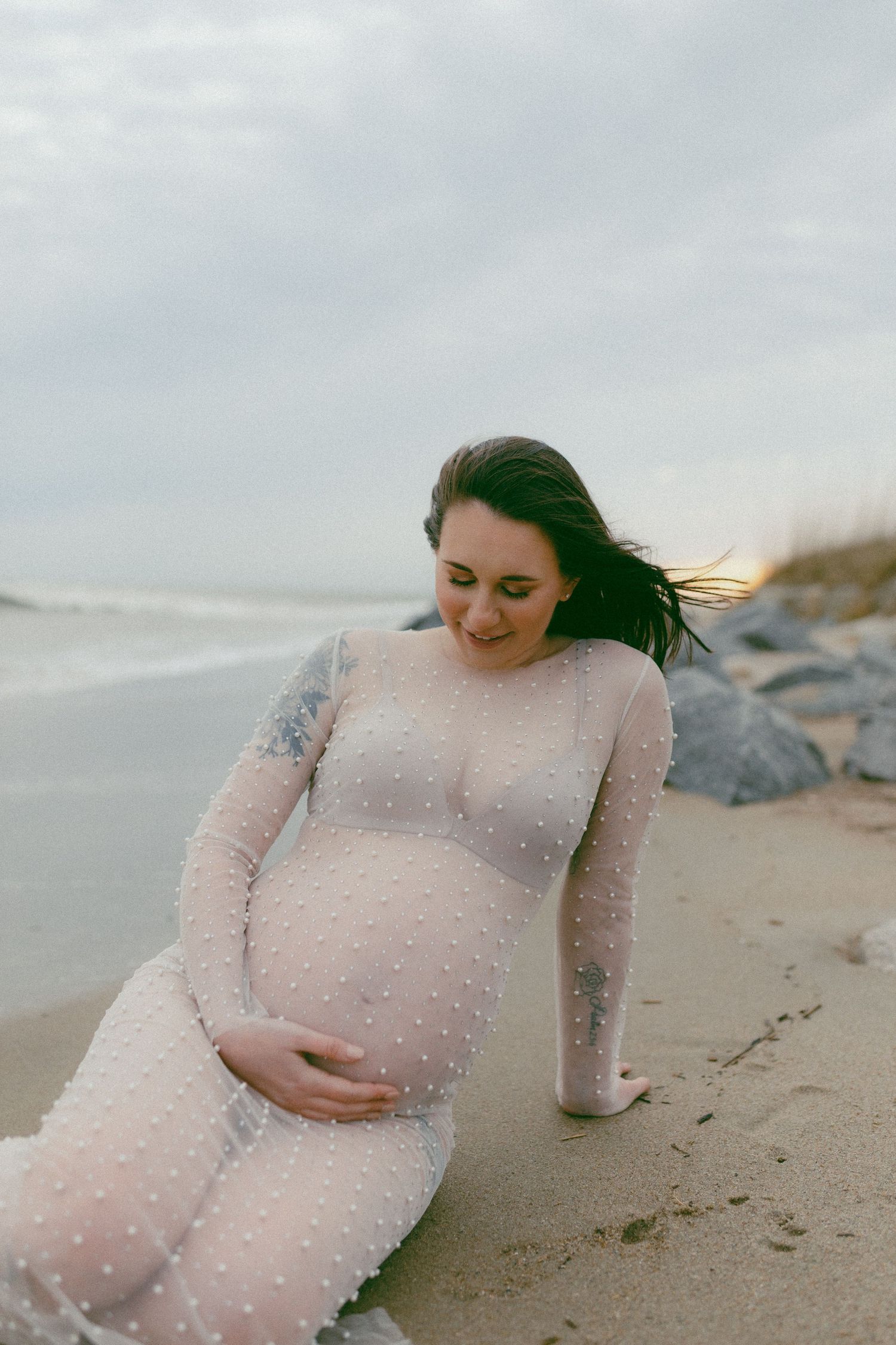 A pregnant woman in a sheer white lace dress poses on a cloudy beach with wind-swept hair.