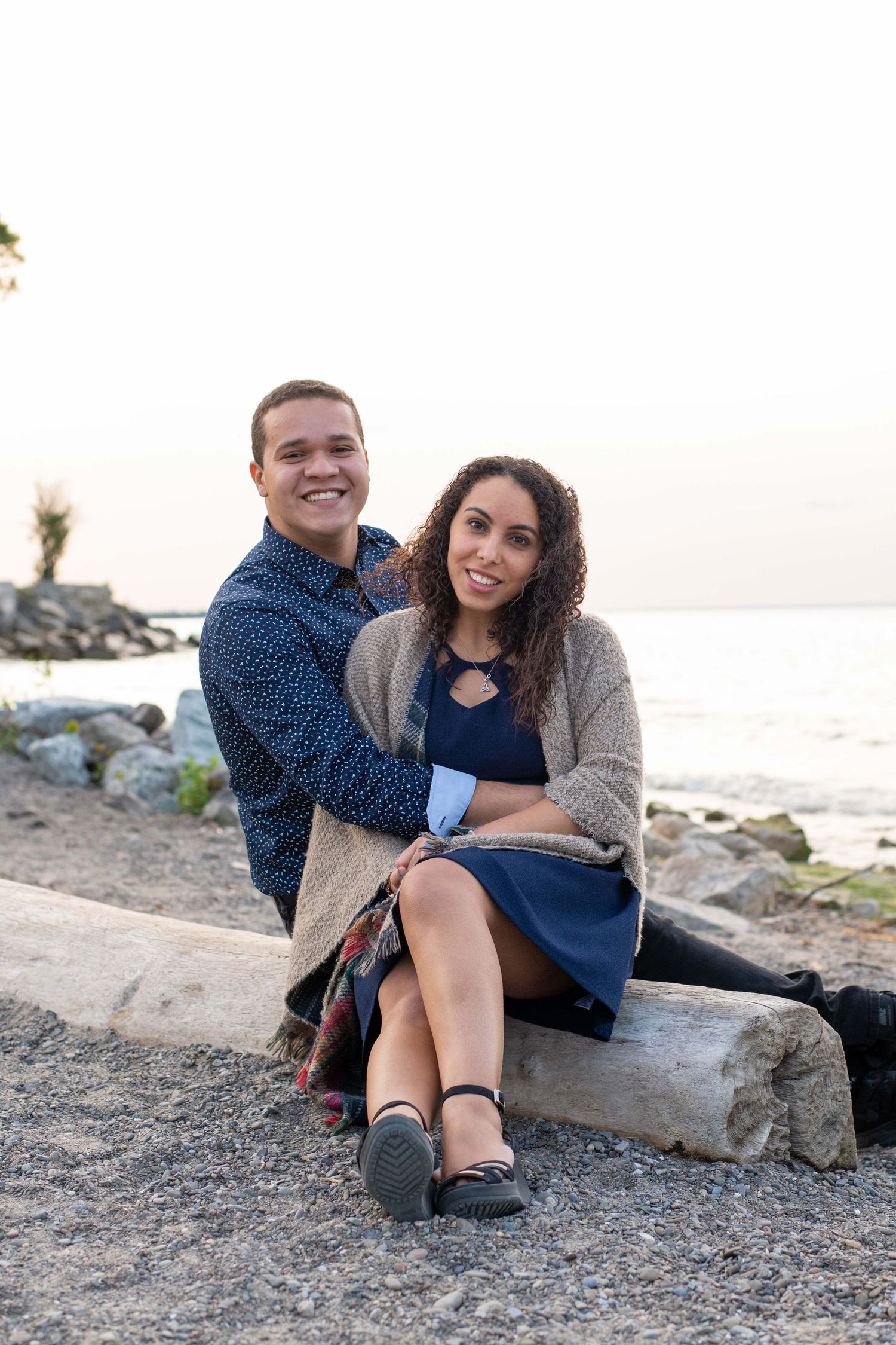 A couple sits together on driftwood along a rocky beach at sunset by the lake.