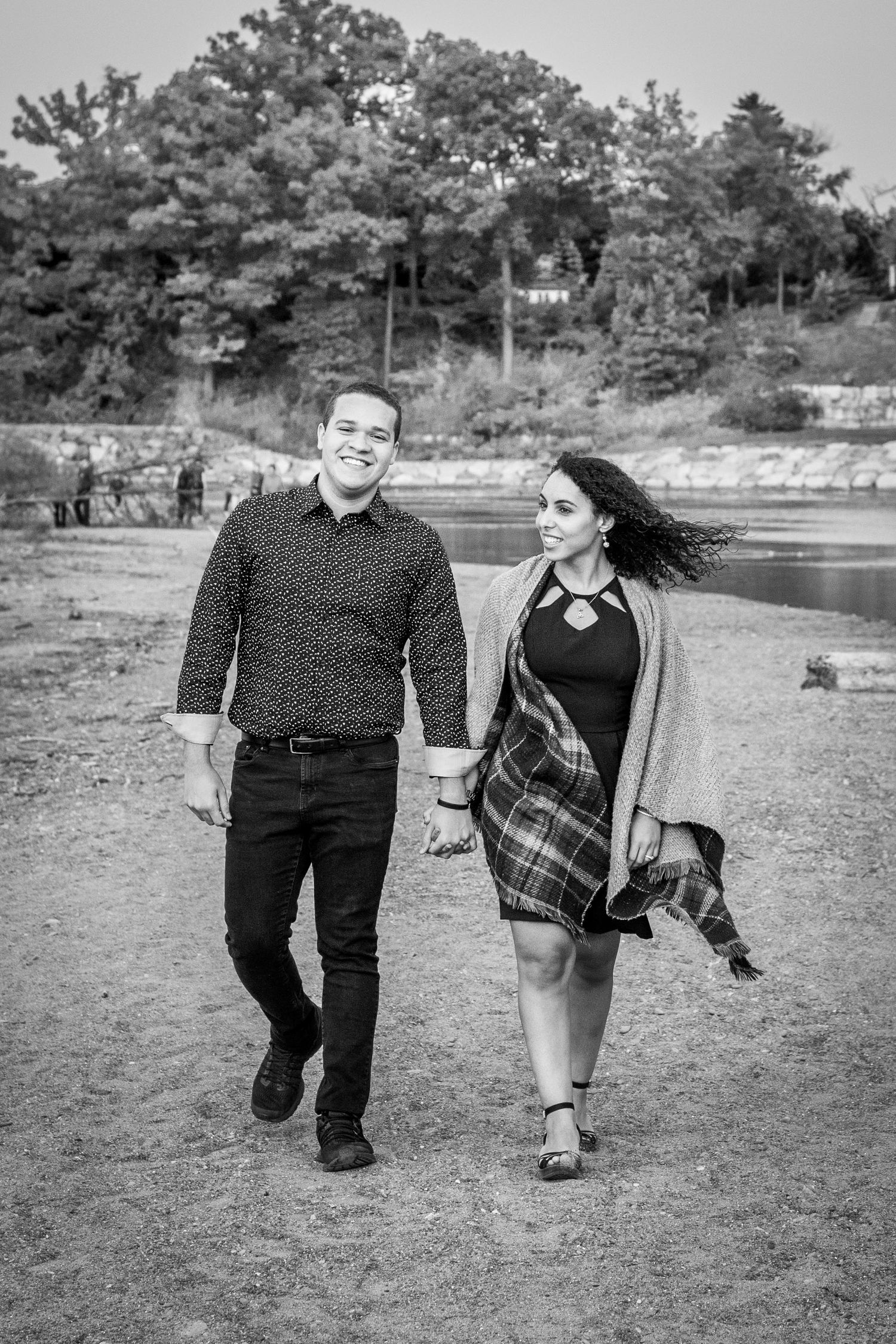 A black and white photo of a couple walking hand in hand on a beach path near trees.
