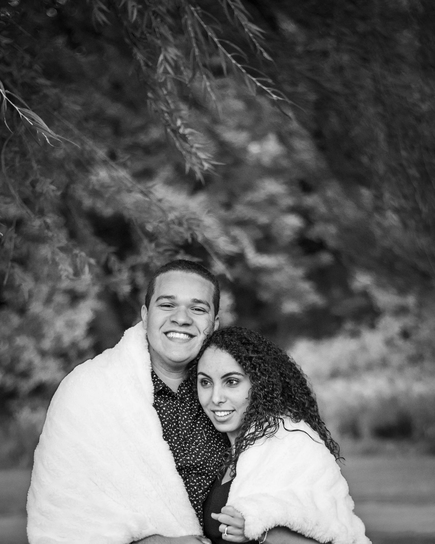 A black and white portrait showing a warm embrace wrapped in cozy blankets outdoors.