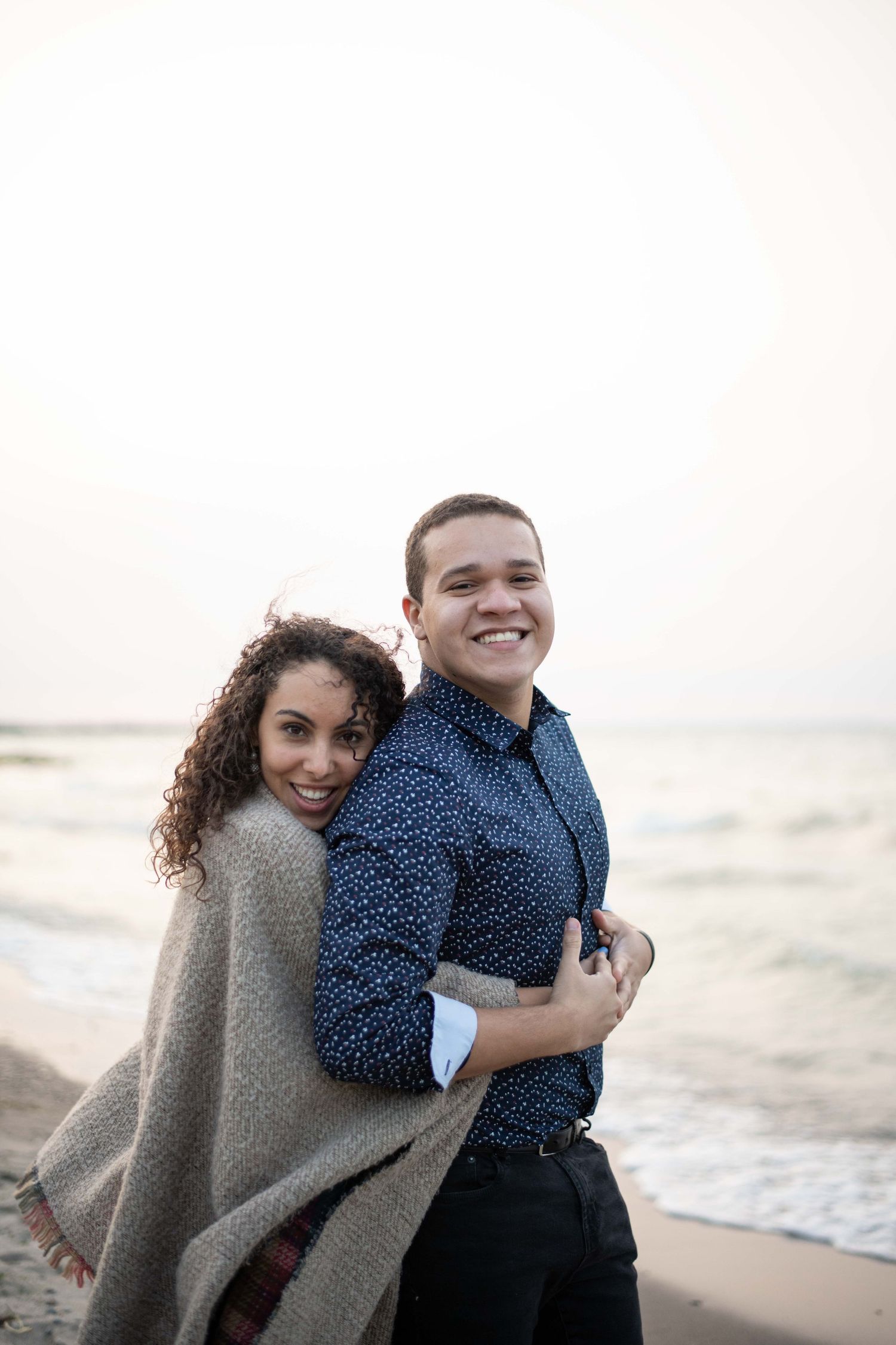 A romantic couple wrapped in a blanket shares a tender moment on the beach at dusk.