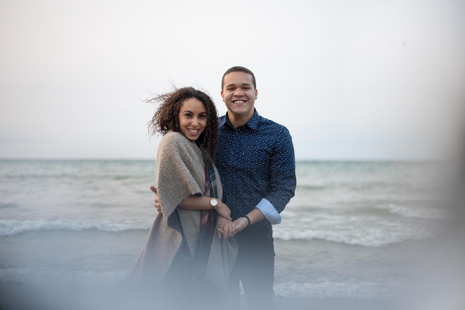 A couple embraces on a beach with waves and overcast sky in the background.