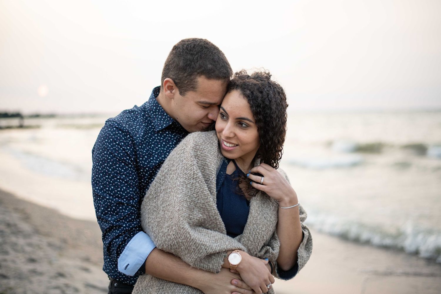 A romantic couple embraces on a peaceful beach at dusk with gentle waves in the background.