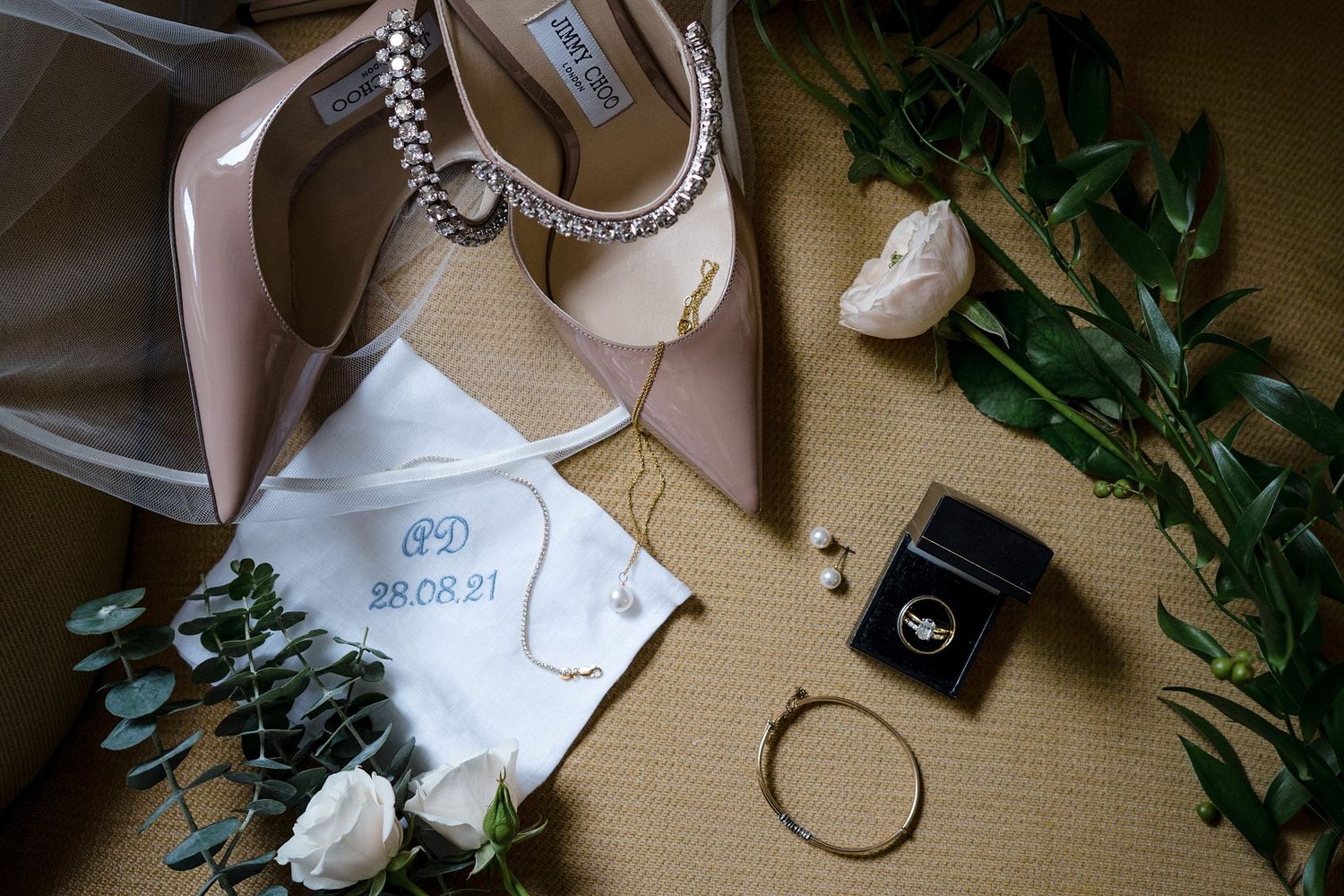 Wedding accessories including Jimmy Choo heels, engagement ring, delicate floral sprigs and wedding stationery arranged on beige surface.