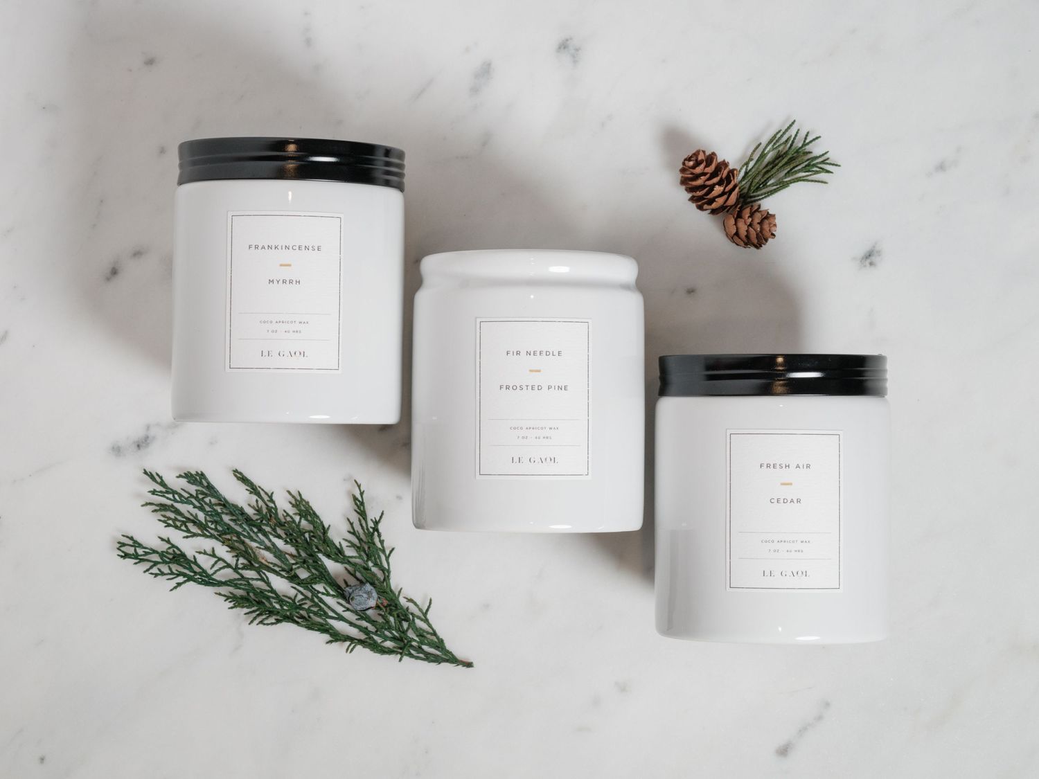 Three white minimalist candle containers with pine sprigs on marble background.