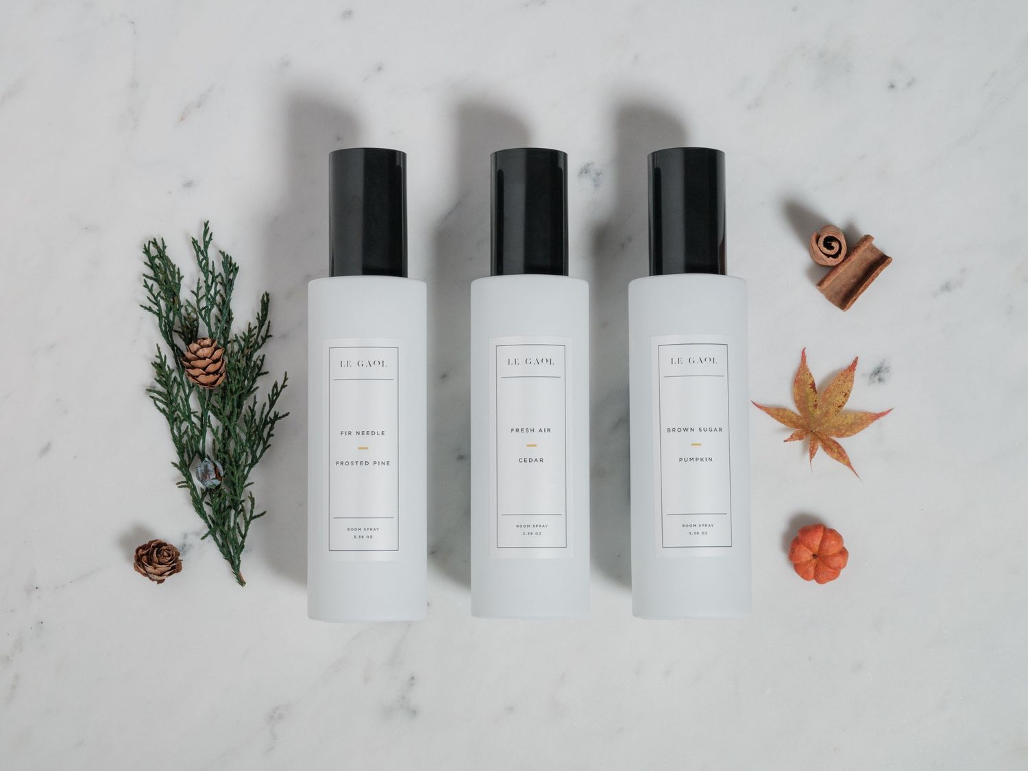Three minimalist white spray bottles arranged with decorative pine sprigs and fall leaves on marble surface.