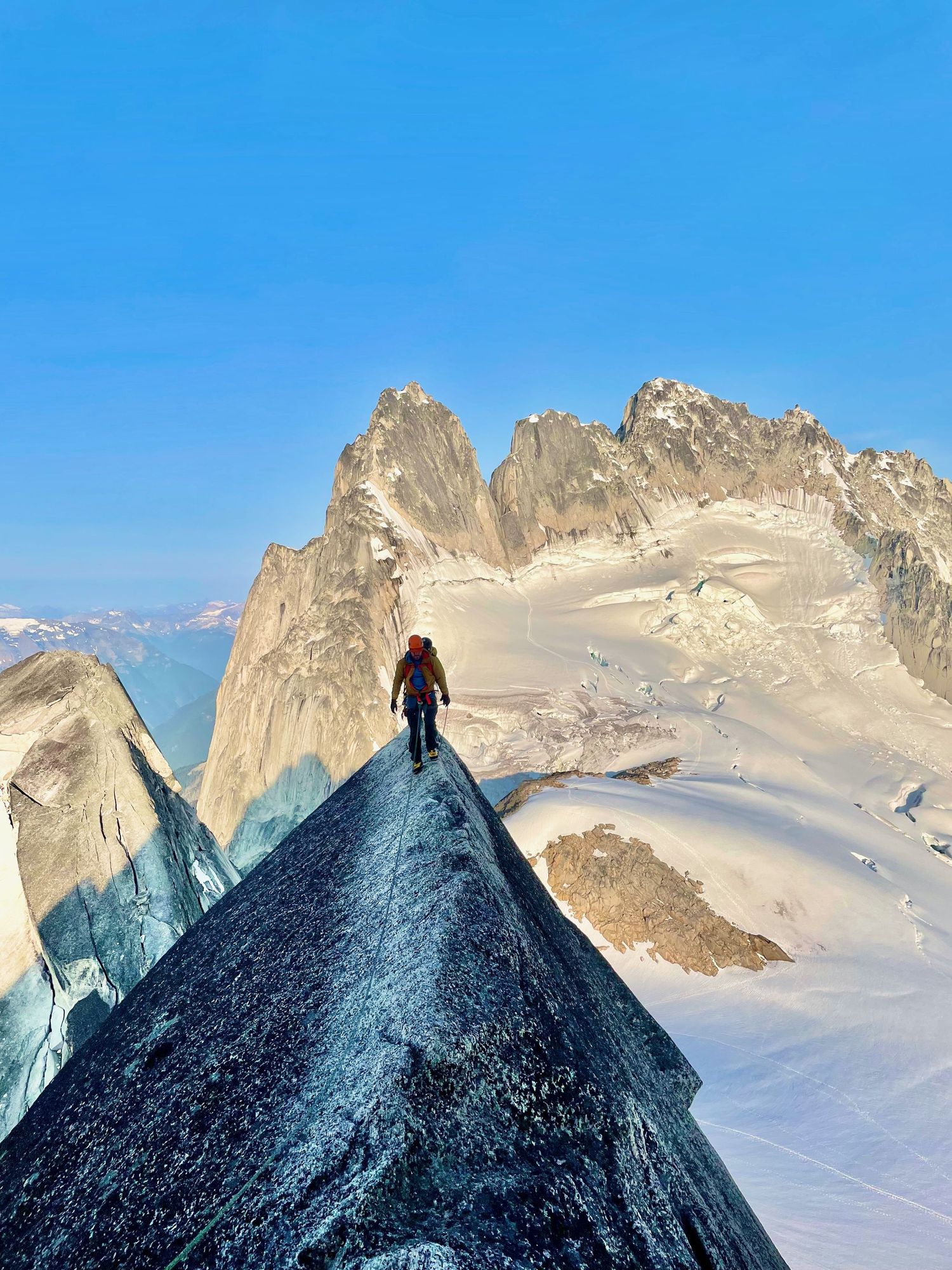 Evan Will stands atop a narrow mountain ridge with dramatic snow-covered peaks and blue sky in the background in Bugaboos Park, Canada