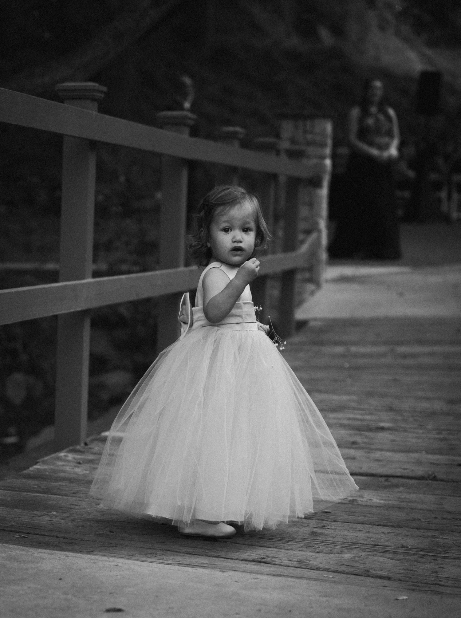 A small child on a wooden boardwalk wearing a flowing white tulle dress stands against a railing in black and white.