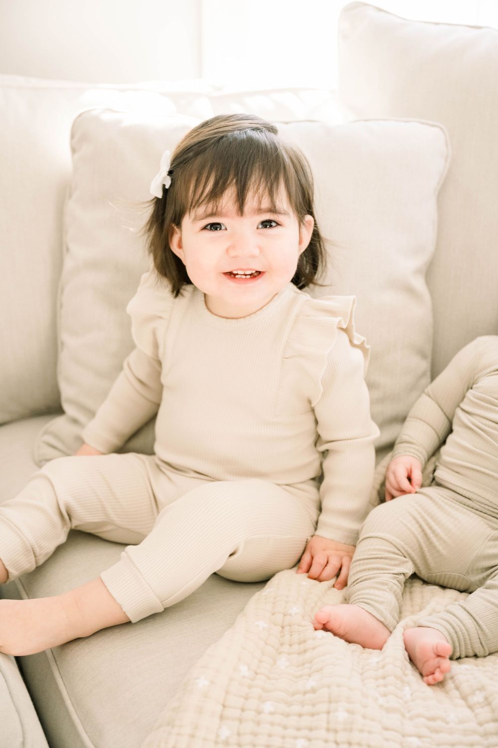 Toddler in beige outfit sitting on light colored couch with decorative cushions.