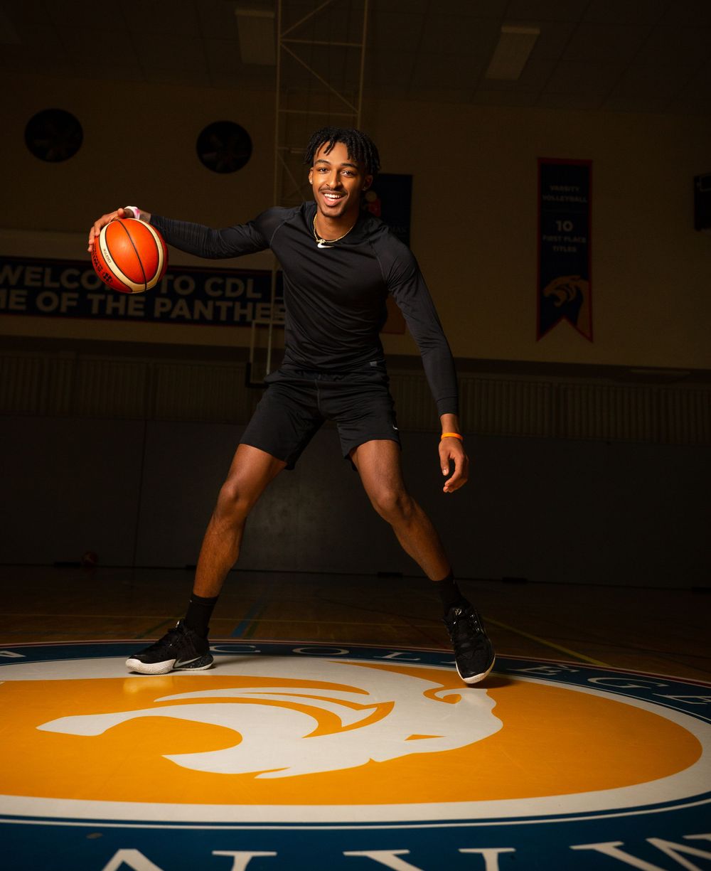 Basketball player dribbling on a gymnasium court with team logo in dramatic lighting.