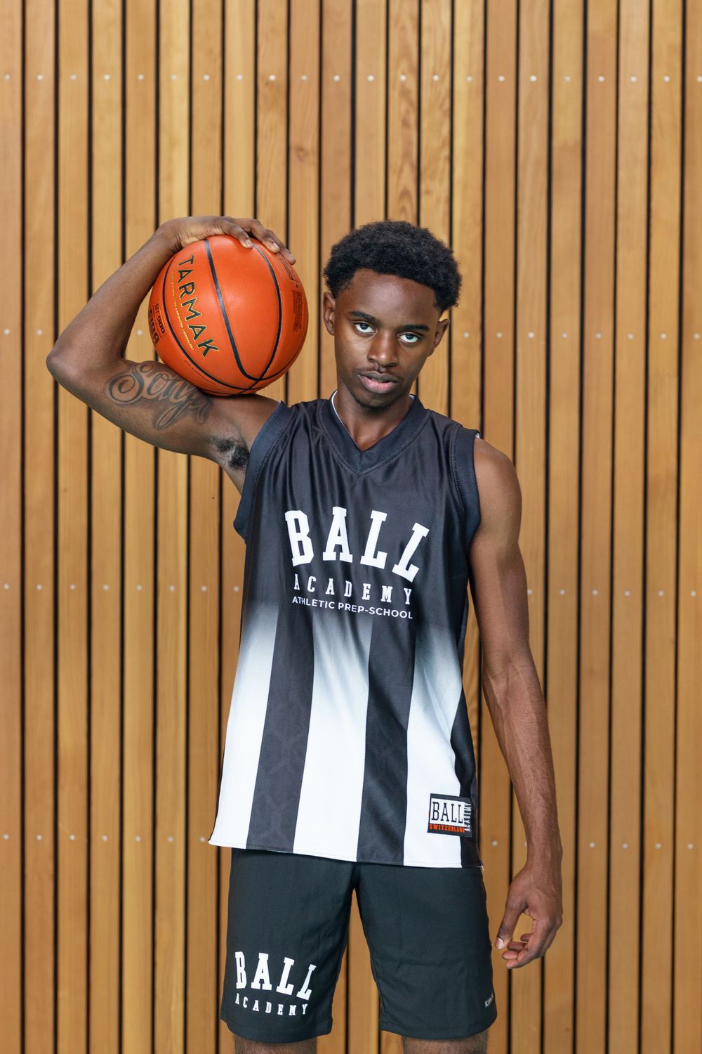 Basketball player in Ball Academy uniform poses with ball against wooden wall background.